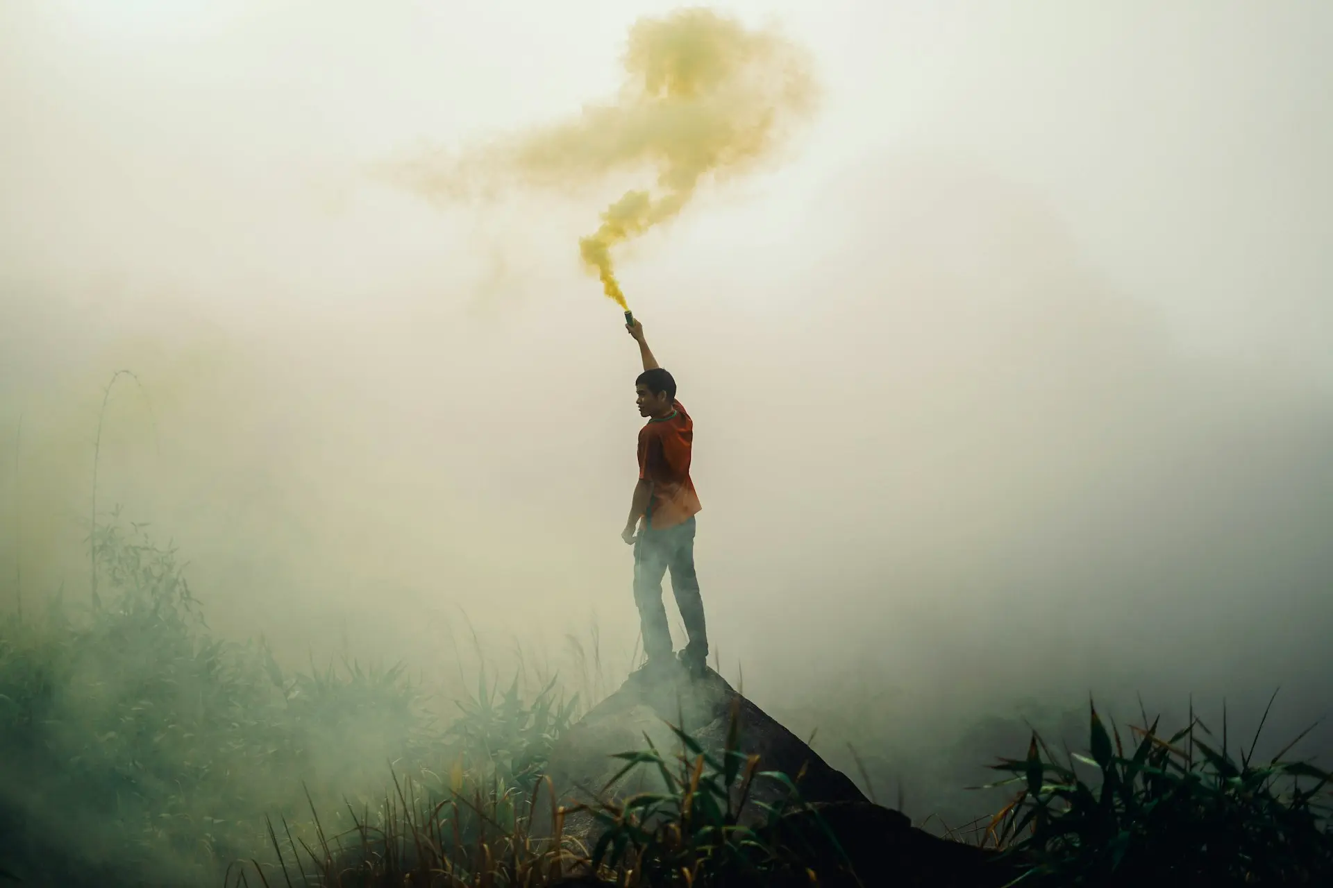 man standing on rock formation holding yellow flare surrounded by fogs